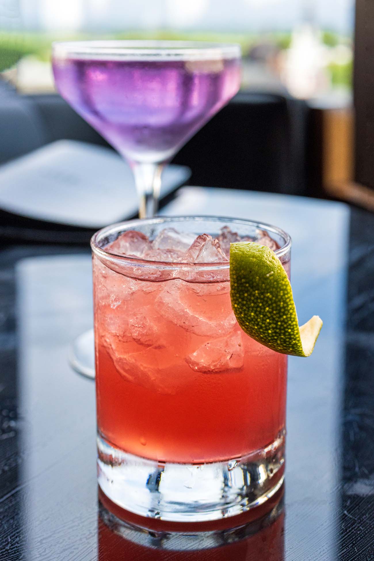he Mezcolada cocktail from The Exchange Lancaster featuring a coral-pink drink in a rocks glass with ice and a lime wheel garnish, with a purple cocktail blurred in the background, both sitting on a dark table.