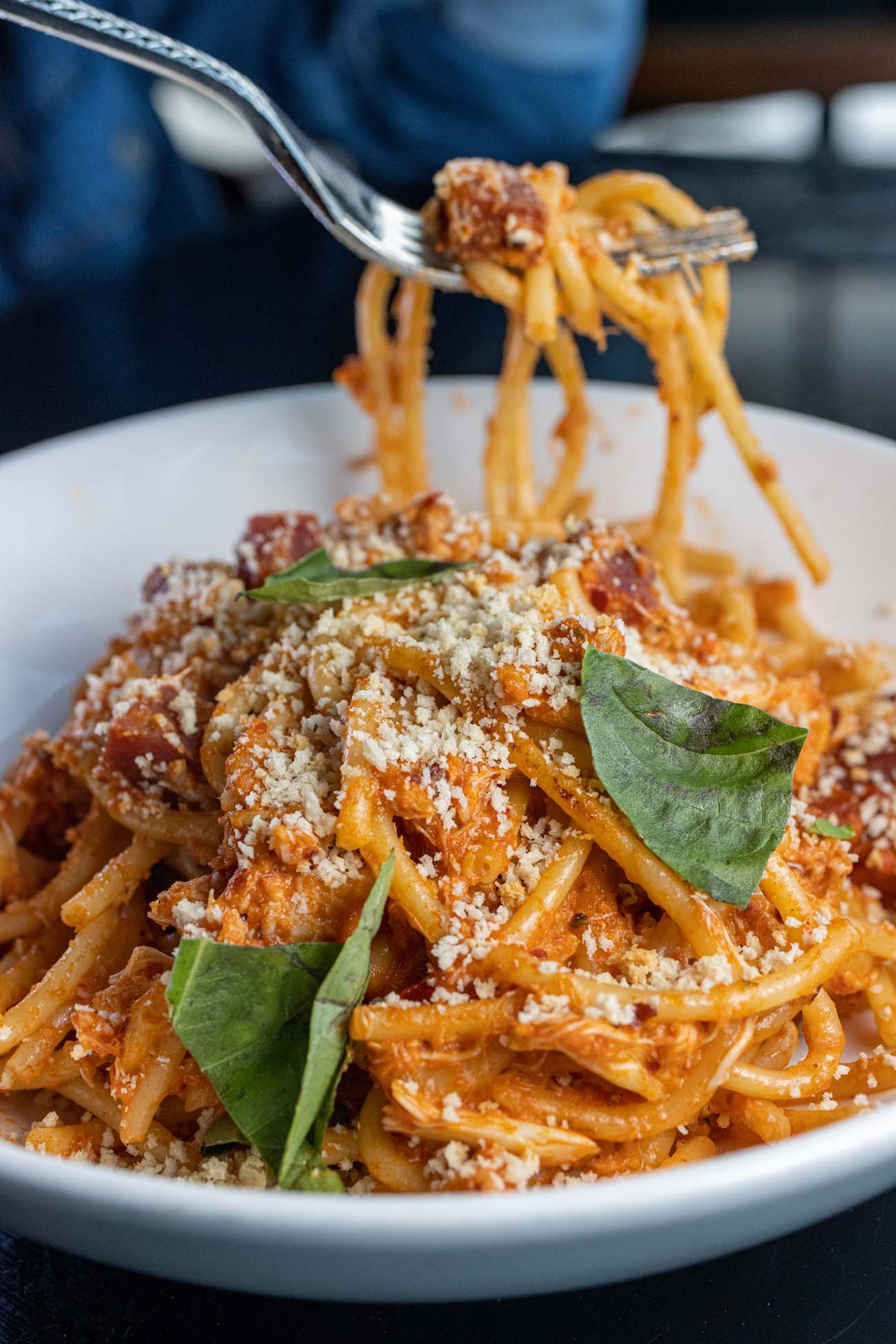 Action shot of the Spicy Crab Bucatini from The Exchange Lancaster showing pasta being twirled on a fork above the white bowl, with thick bucatini noodles in rich tomato sauce topped with grated cheese and fresh basil leaves.
