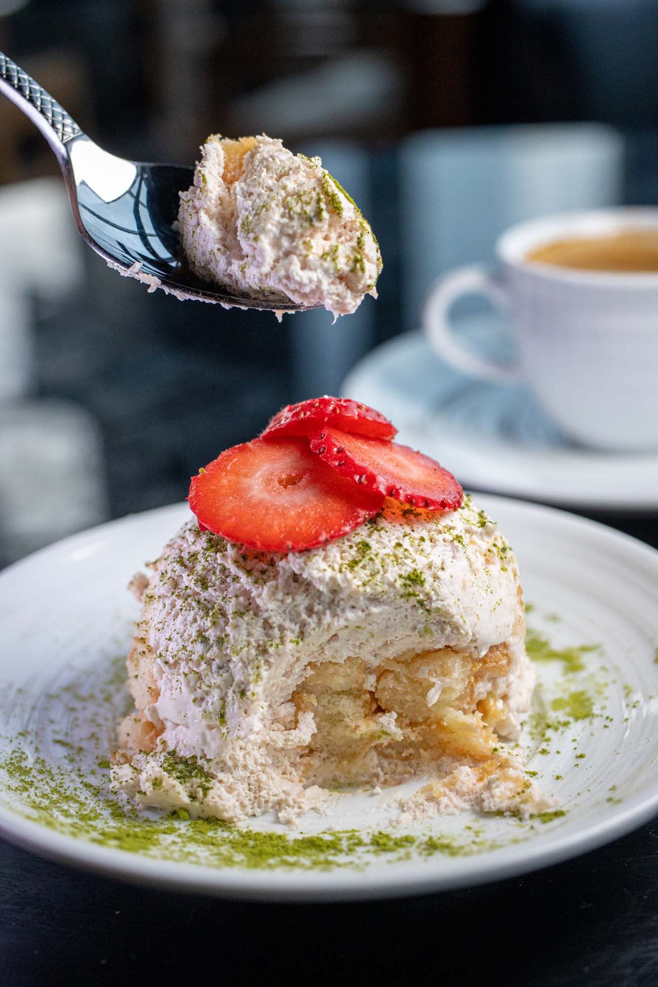 Strawberry Matcha Tiramisu from The Exchange Lancaster on a white plate dusted with green matcha powder, topped with fresh strawberry slices, shown with a spoonful being lifted to reveal the layered cream and cake interior, with a coffee cup blurred in the background.