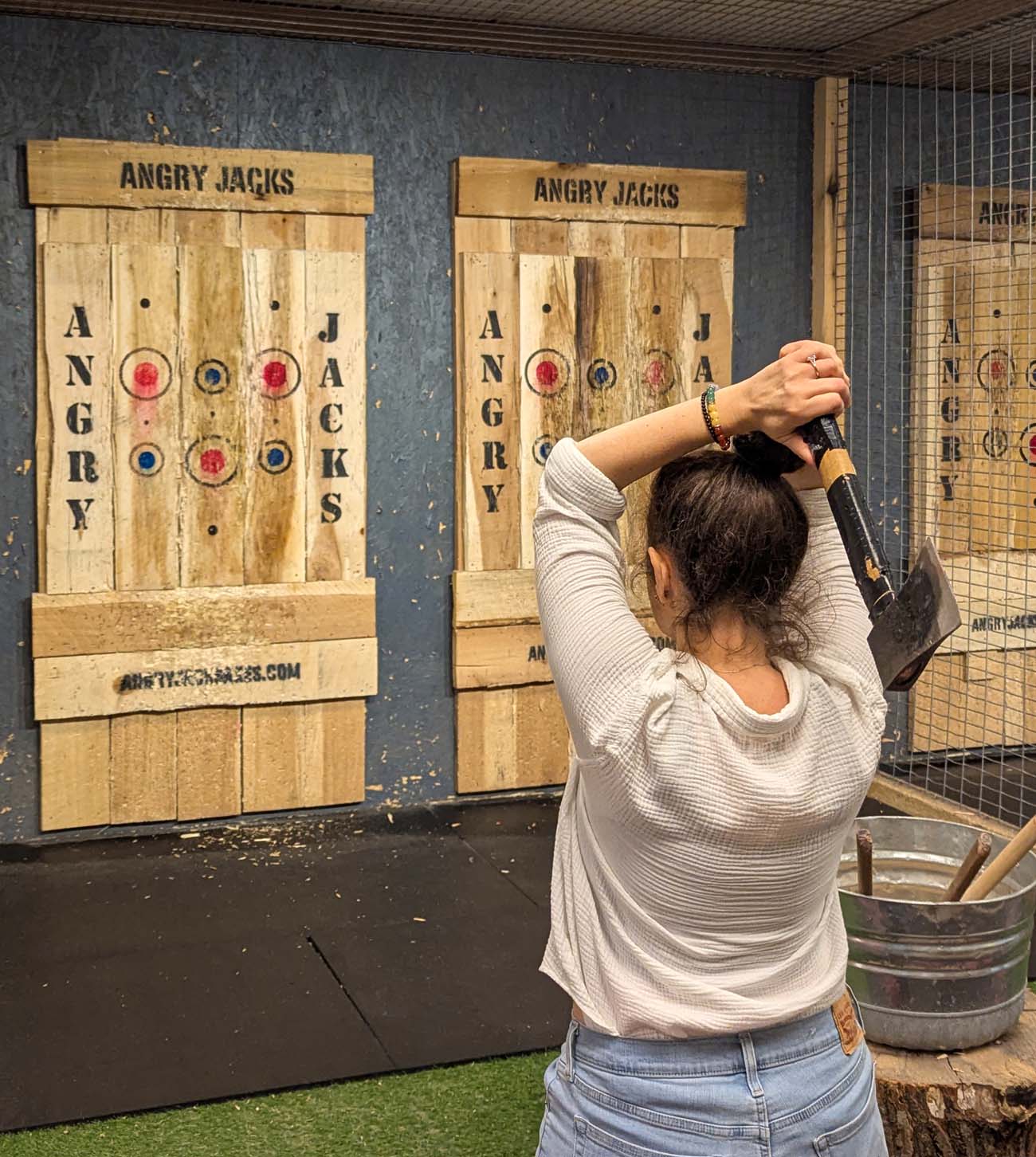 Heather from Better Living throwing axe at wooden Angry Jacks target boards in industrial-style axe throwing venue with metal cage safety barriers
