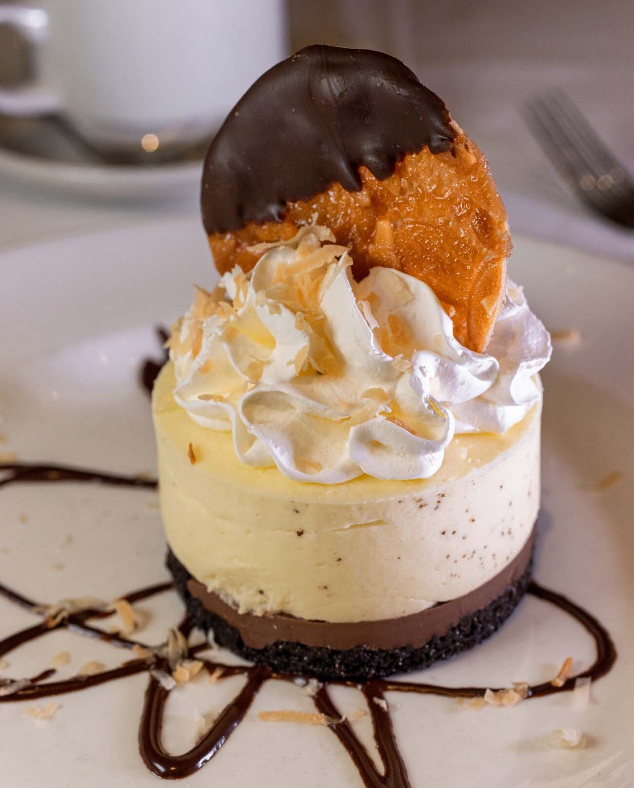 Elegant coconut cream pie with chocolate-dipped tuile, whipped cream, and toasted coconut on white plate with chocolate sauce drizzle and coffee cup in background