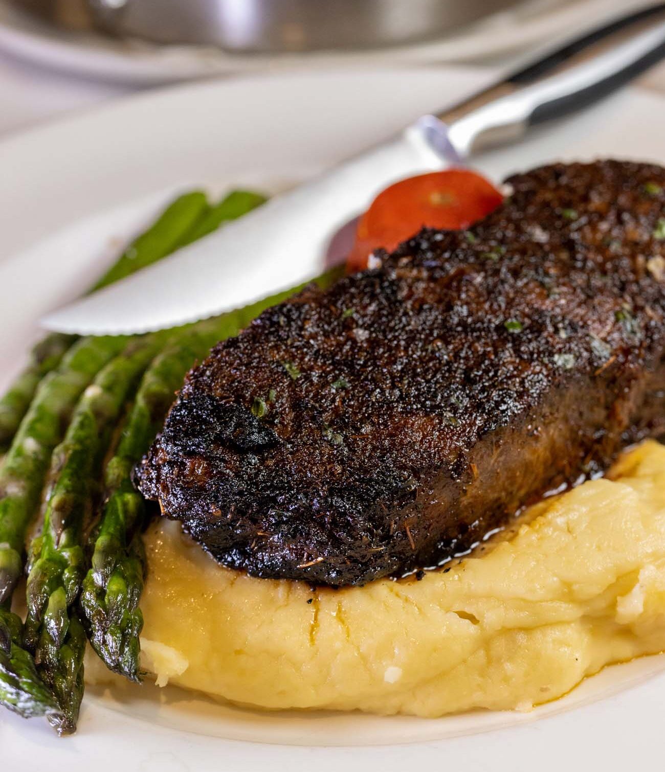 Perfectly seared New York strip steak with whipped potatoes and asparagus on white plate, with sauté pan visible in background at upscale steakhouse