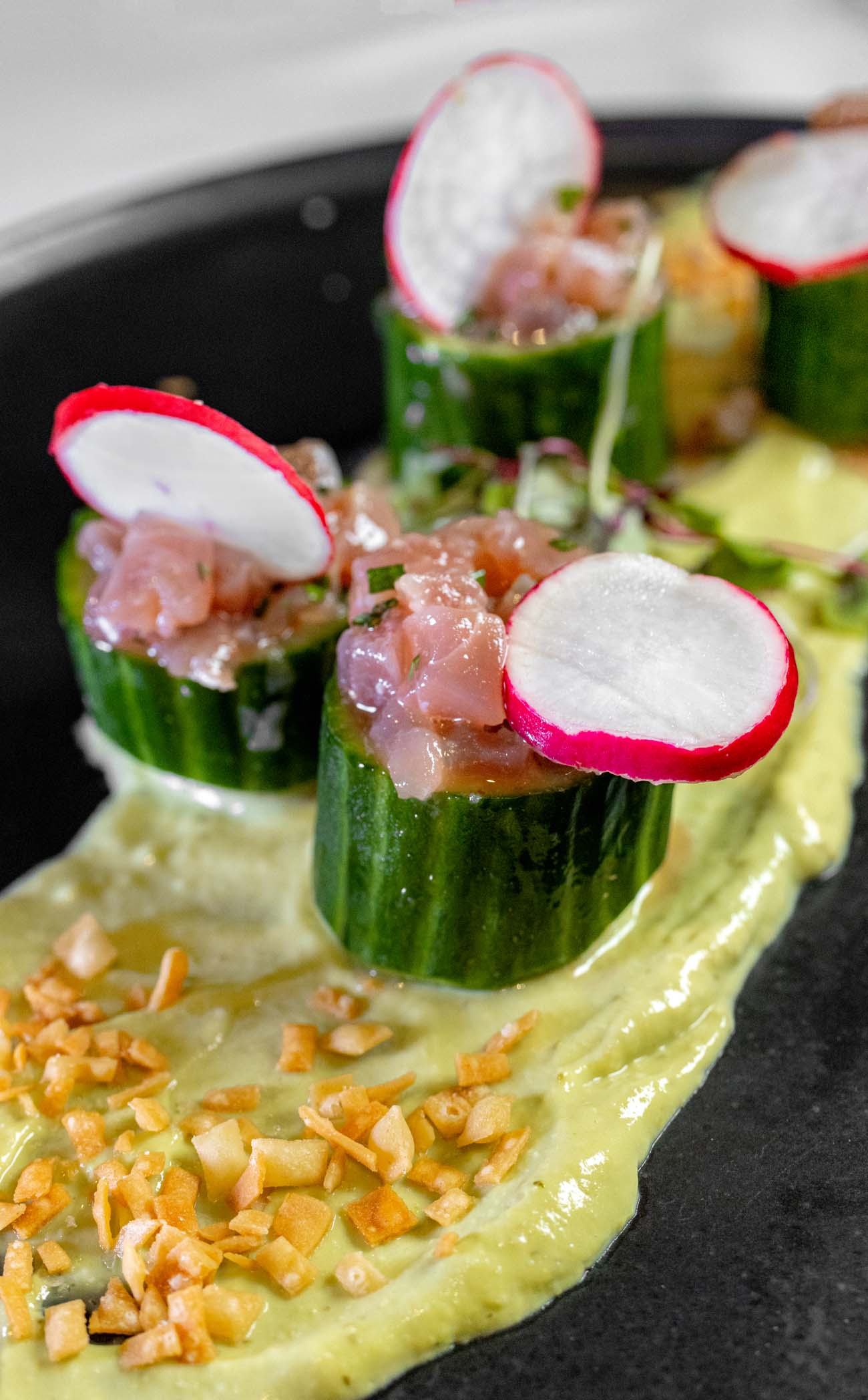 Elegant ahi tuna tartare served in hollowed cucumber cups with radish garnish and sesame seeds on black slate plate with avocado sauce