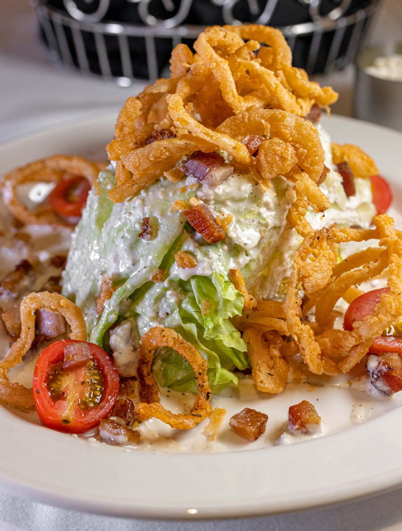 Generous wedge salad topped with crispy fried onion strings, bacon bits, blue cheese dressing and cherry tomatoes on white plate with decorative silver basket
