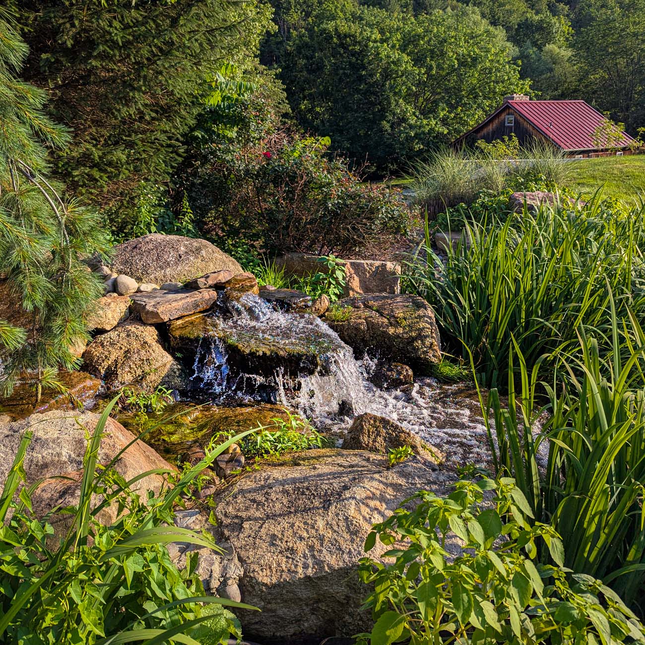 A cascading water feature built into natural stone formations at Glasbern Inn, surrounded by lush green grasses and plantings, with a red-roofed building visible in the wooded background