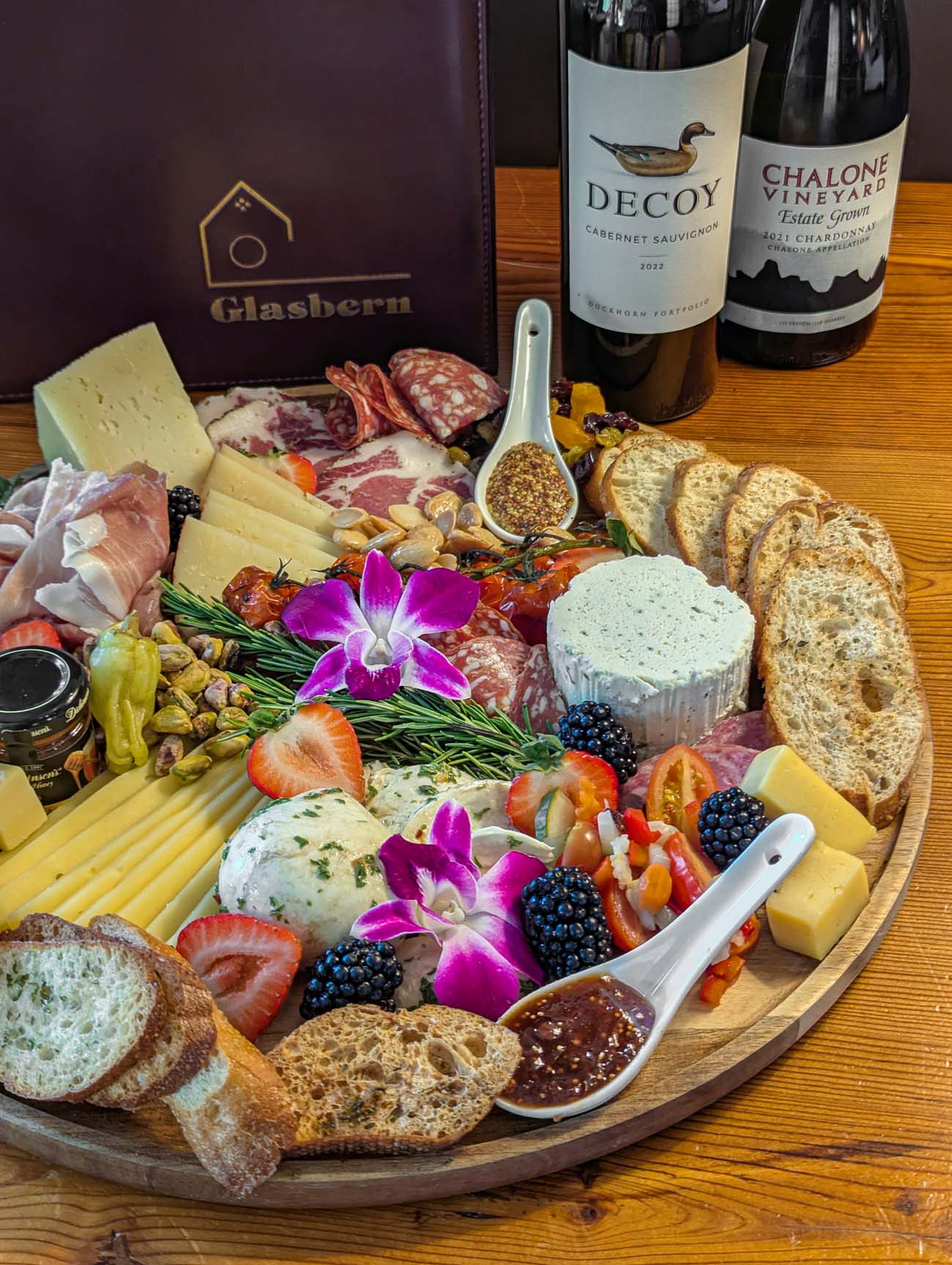 laborate charcuterie board featuring artisanal meats, aged cheeses, fresh fruits, crackers, and purple orchid garnishes, accompanied by wine bottles and Glasbern branded packaging on a wooden table