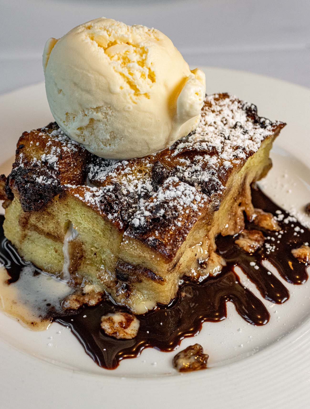 Rectangular portion of golden-brown bread pudding topped with a scoop of vanilla ice cream, drizzled with dark chocolate sauce and garnished with chopped nuts on a white plate