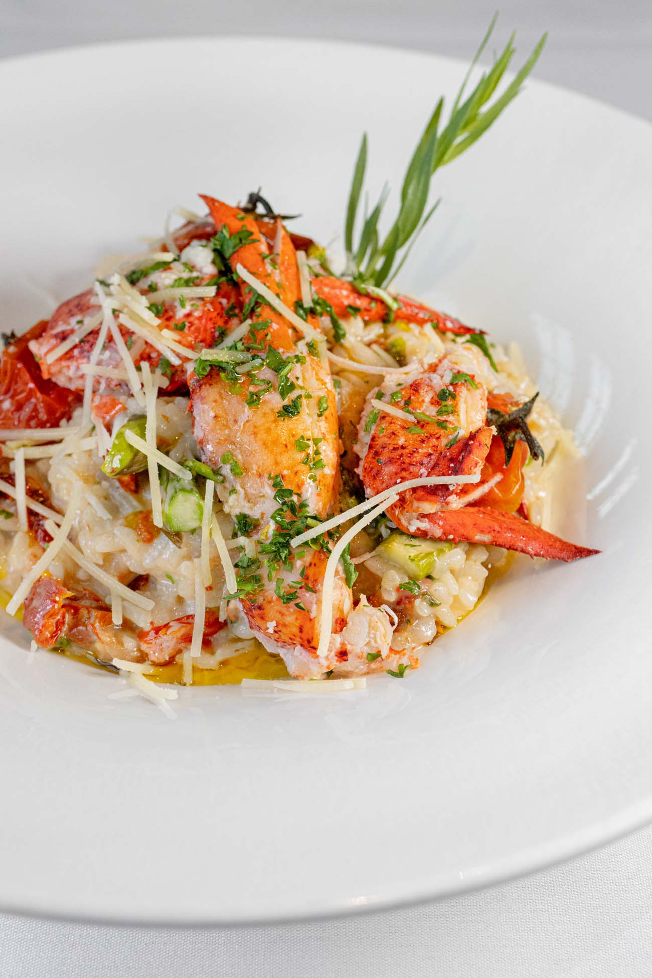 Creamy risotto topped with generous chunks of bright red lobster meat, fresh asparagus spears, sun-dried tomatoes, and fresh herbs, garnished with a sprig of rosemary on a white plate