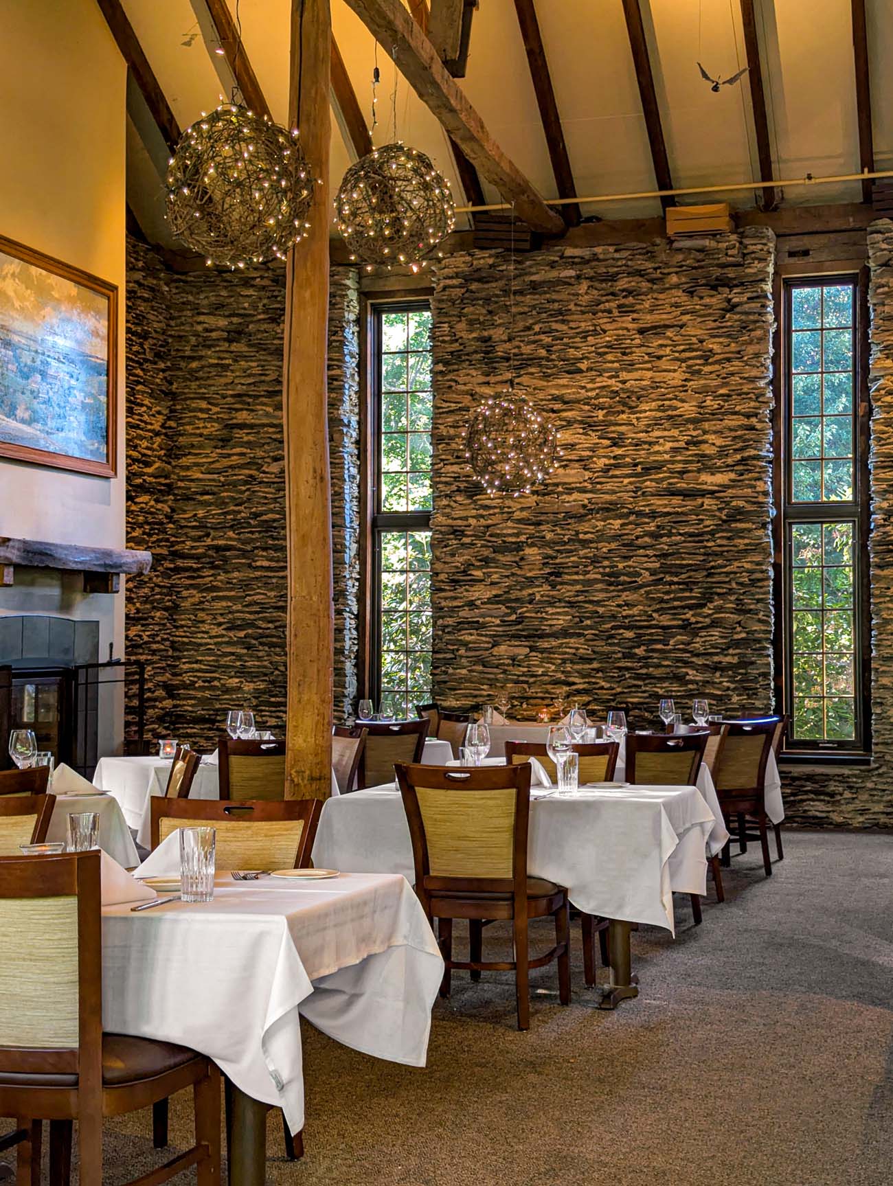 Sophisticated restaurant interior featuring exposed stone walls, tall windows, rustic timber beams, modern pendant lighting, and tables set with white linens and wine glasses