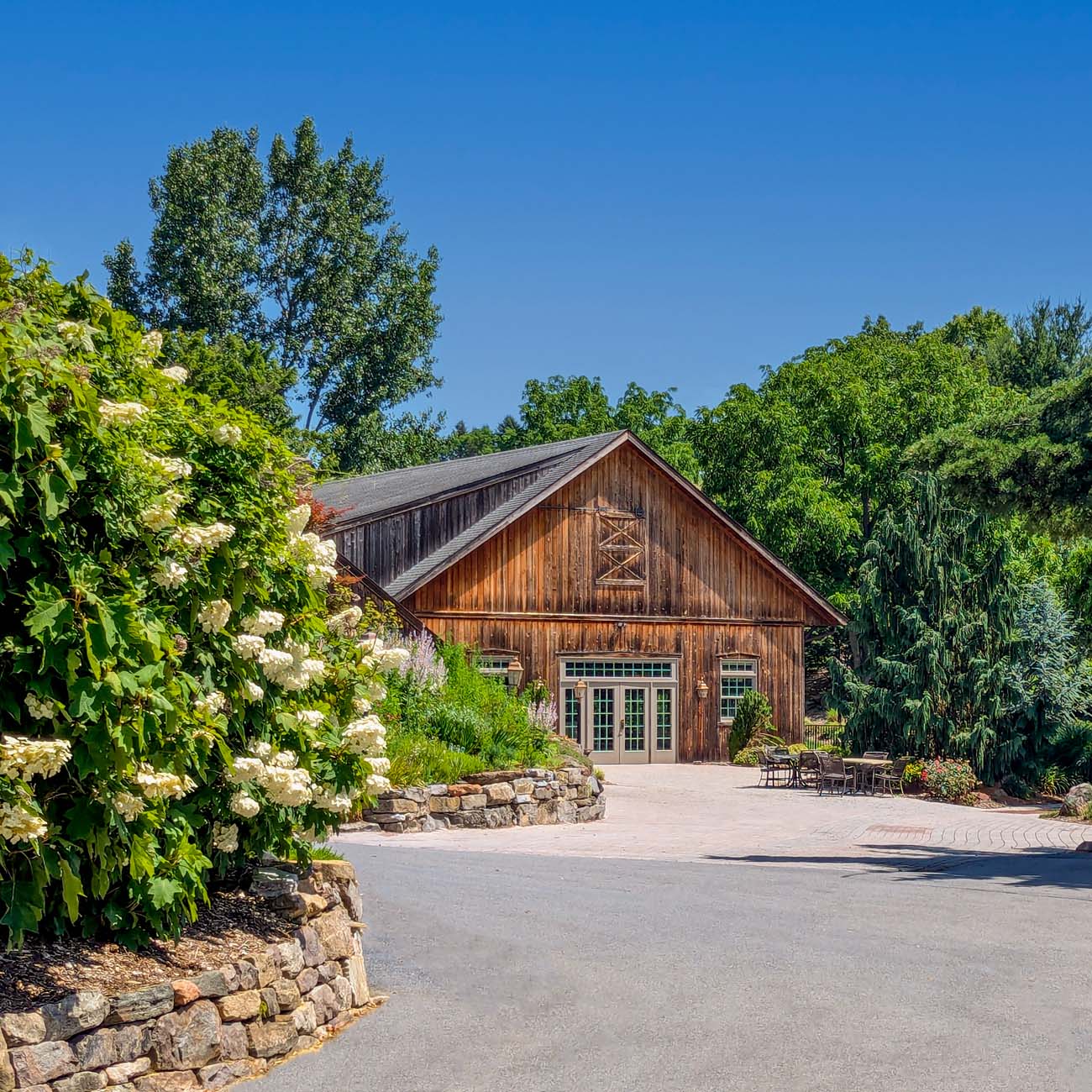 Large rustic wooden building with red metal roof nestled among mature trees, featuring manicured lawns and natural stone landscaping elements in the foreground
