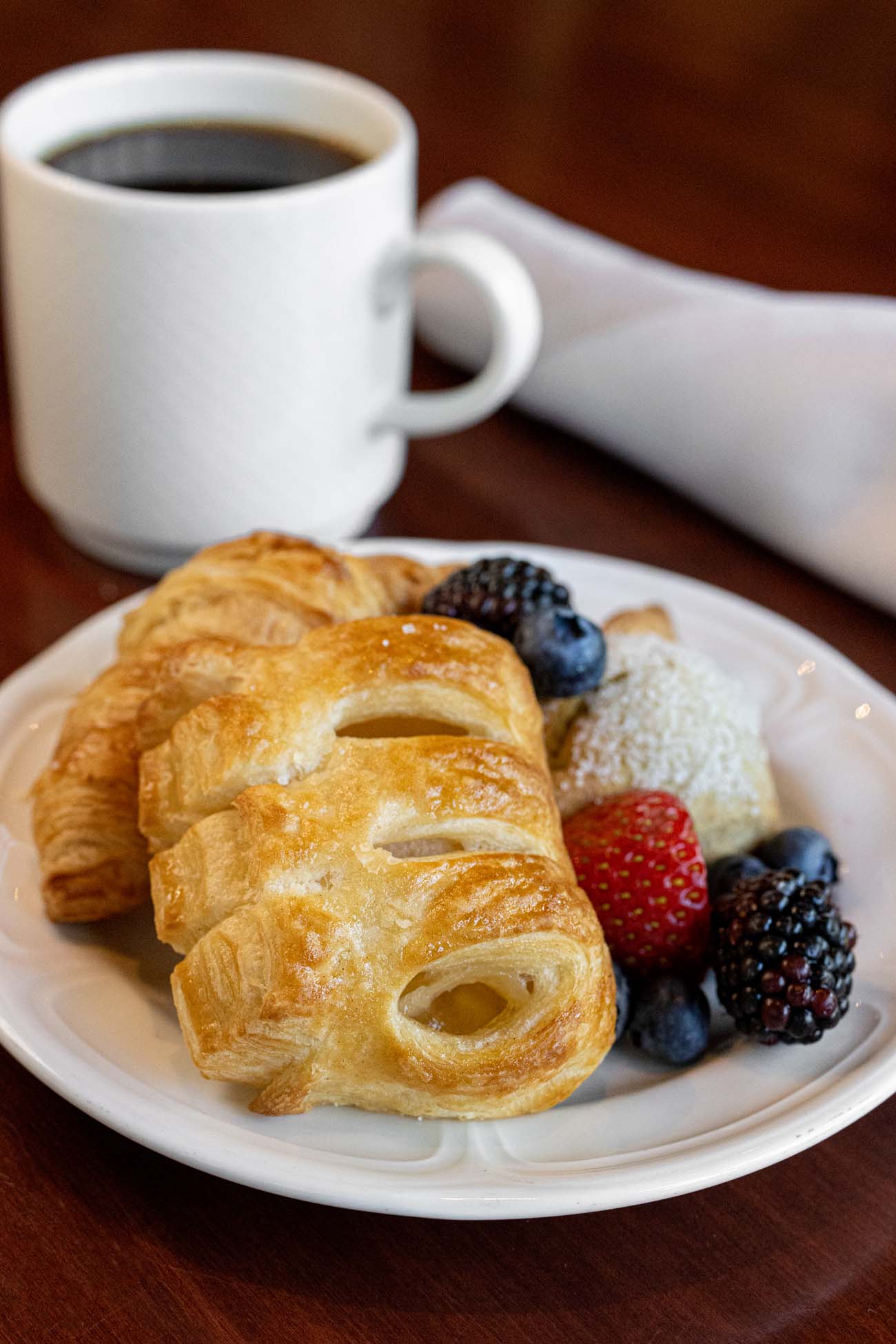 White plate containing golden flaky pastries, fresh berries including strawberries, blackberries and blueberries, alongside a white coffee cup on a dark wooden surface