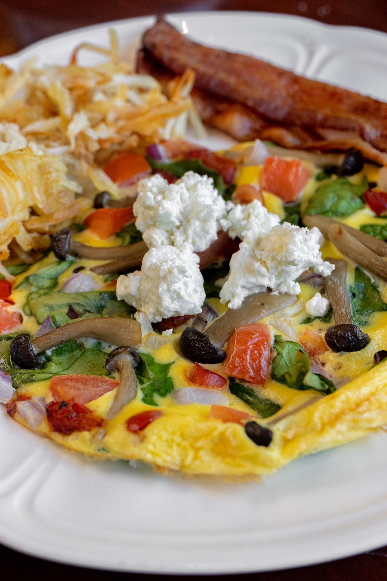 Colorful frittata filled with red peppers, spinach, mushrooms, onions and topped with white goat cheese crumbles, served with breakfast sausage and hash browns on a white plate