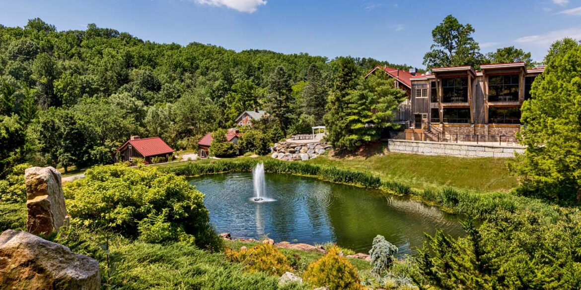 Panoramic view of Glasbern's main pond and buildings. Elevated view showing a large pond with central fountain surrounded by landscaped grounds, with multiple rustic buildings including barns and guest accommodations scattered throughout the wooded property