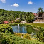 Panoramic view of Glasbern's main pond and buildings. Elevated view showing a large pond with central fountain surrounded by landscaped grounds, with multiple rustic buildings including barns and guest accommodations scattered throughout the wooded property