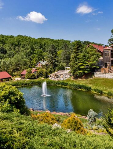Panoramic view of Glasbern's main pond and buildings. Elevated view showing a large pond with central fountain surrounded by landscaped grounds, with multiple rustic buildings including barns and guest accommodations scattered throughout the wooded property