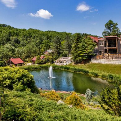 Panoramic view of Glasbern's main pond and buildings. Elevated view showing a large pond with central fountain surrounded by landscaped grounds, with multiple rustic buildings including barns and guest accommodations scattered throughout the wooded property