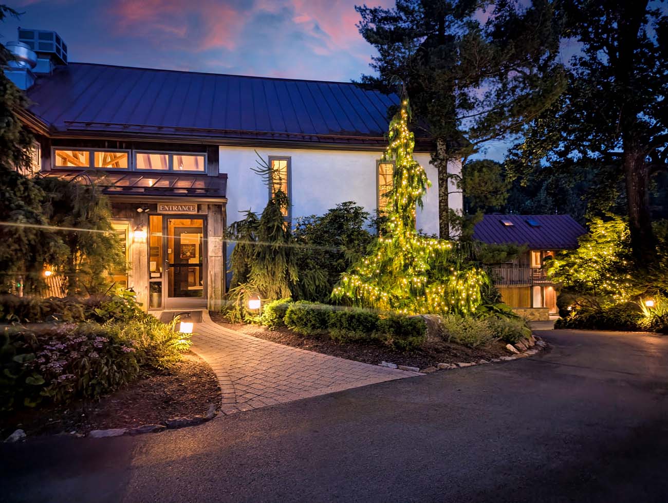 Historic white building with multiple windows glowing warmly in the evening light, featuring an illuminated entrance portico and surrounded by mature trees and landscaping