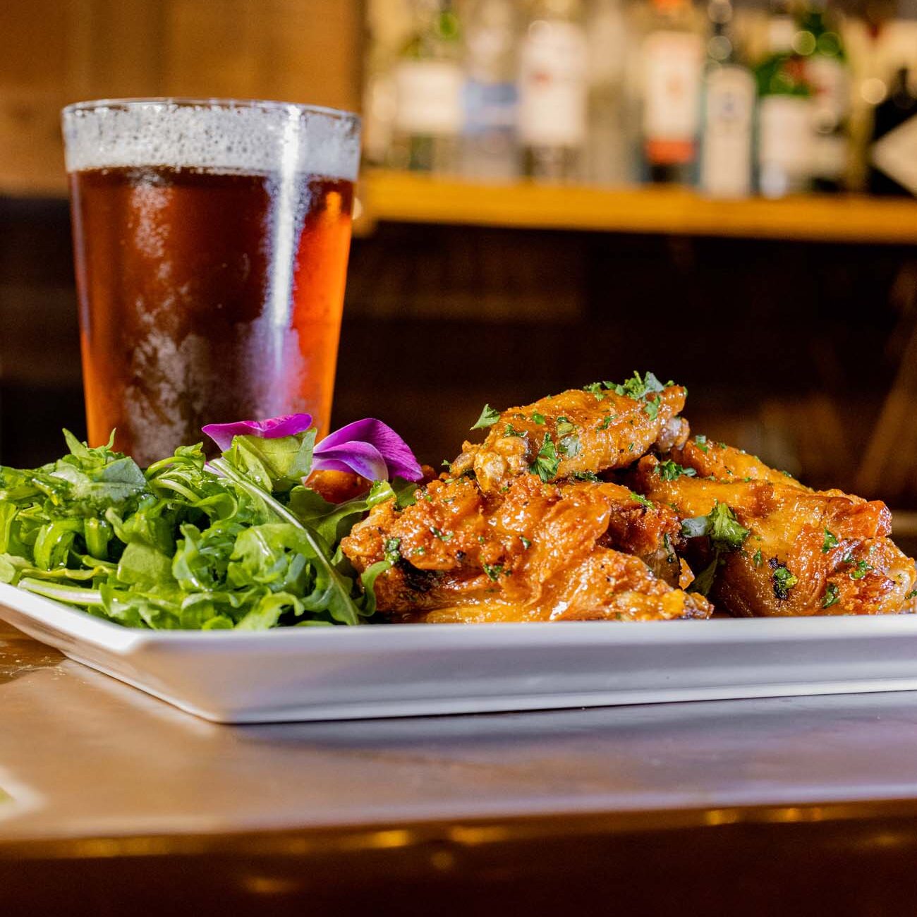 Plate of glazed chicken wings with fresh greens, accompanied by a frothy pint of amber beer on a wooden bar surface with bottles visible in the blurred background