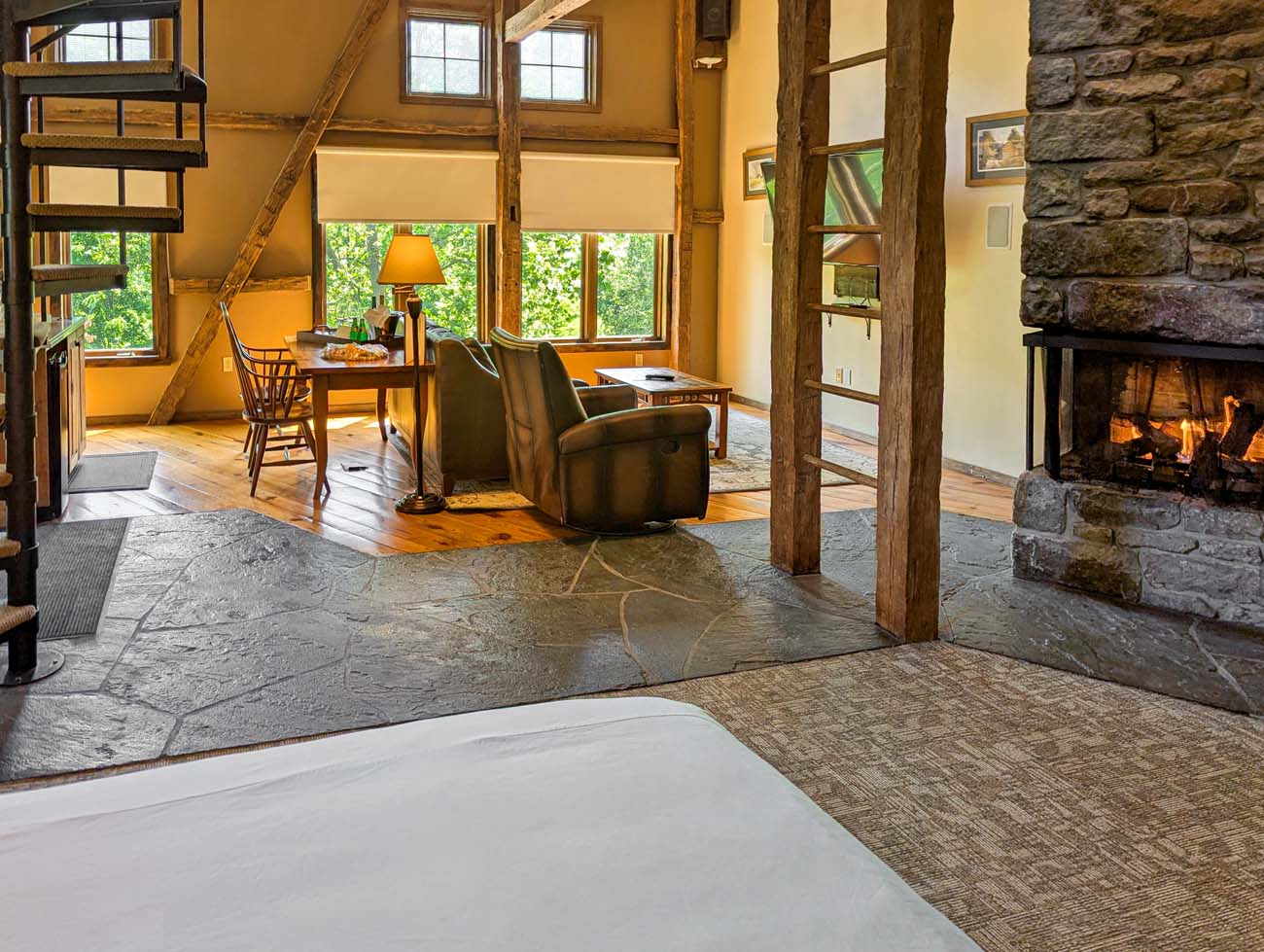 Rustic-luxury suite interior with flagstone flooring, exposed timber framing, stone fireplace, leather seating, and a spiral staircase leading to the loft hot tub area