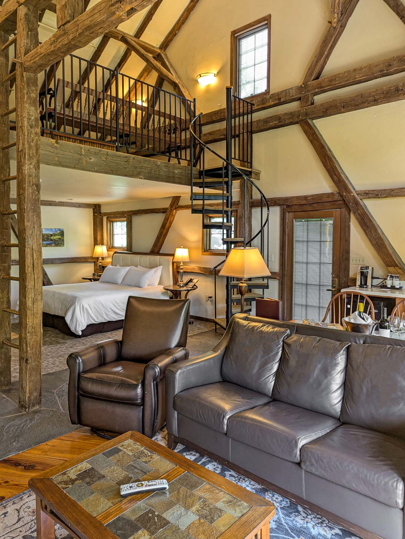 Hotel suite bedroom with white linens and exposed beam ceiling, featuring a dramatic black spiral staircase ascending to an upper level loft area with the hot tub