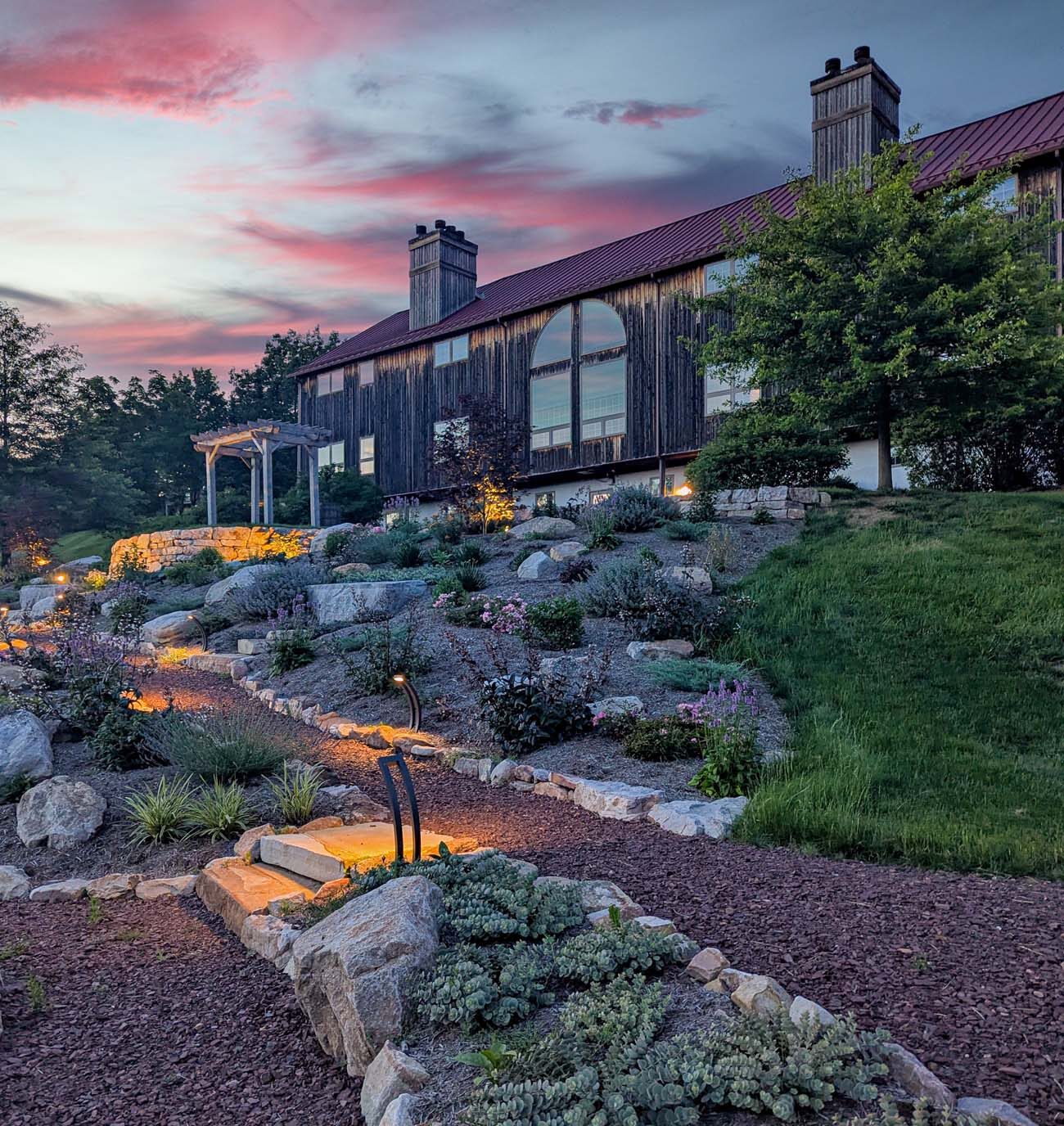 Rustic barn building at dusk with warm interior lighting, surrounded by professionally lit terraced gardens featuring native plants, stone pathways, and artistic landscape lighting under a colorful sunset sky