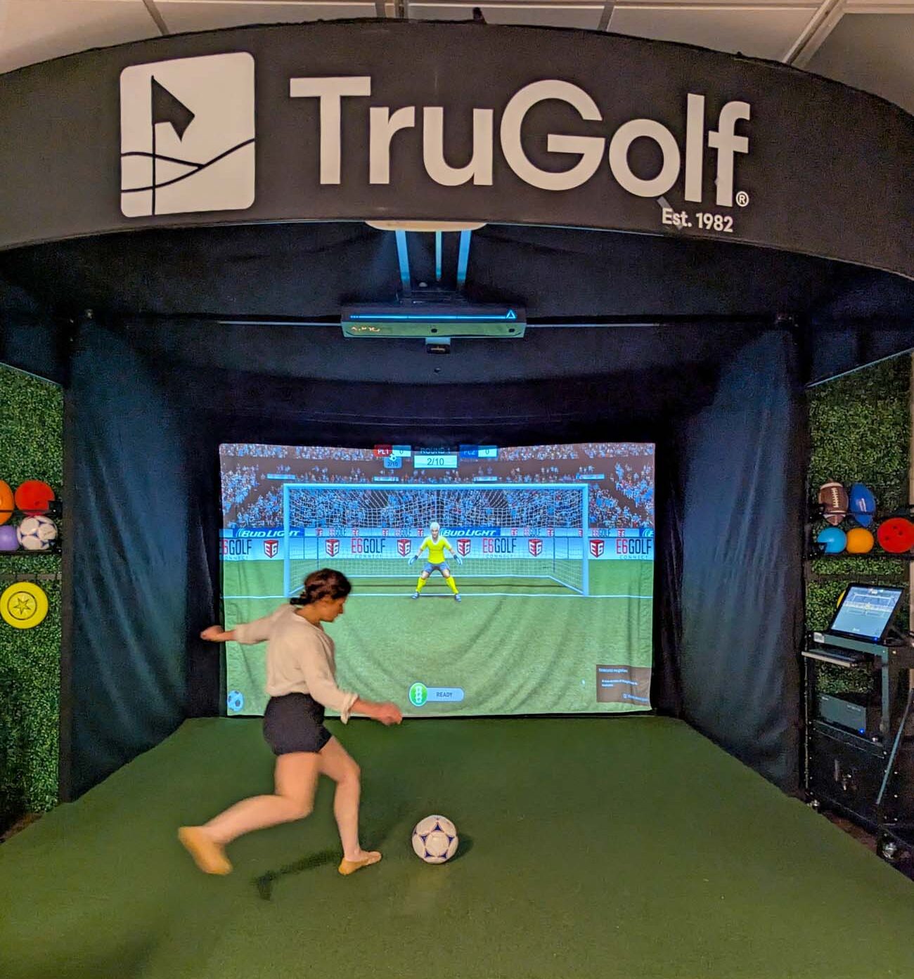 Heather from Better Living kicking soccer ball toward large TruGolf simulator screen showing virtual soccer field and goalkeeper in modern entertainment venue
