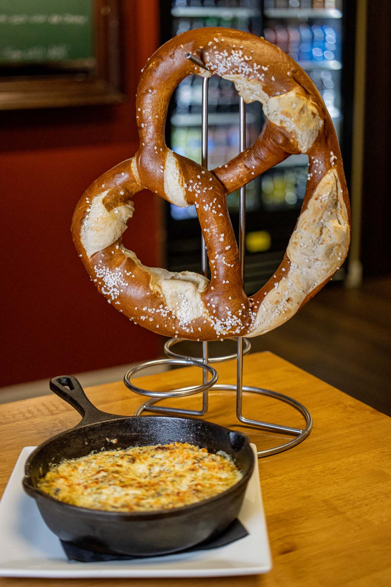 Large warm pretzel mounted on metal stand above cast iron skillet of bubbling spinach artichoke dip on wooden table at Twisted Tees sports bar