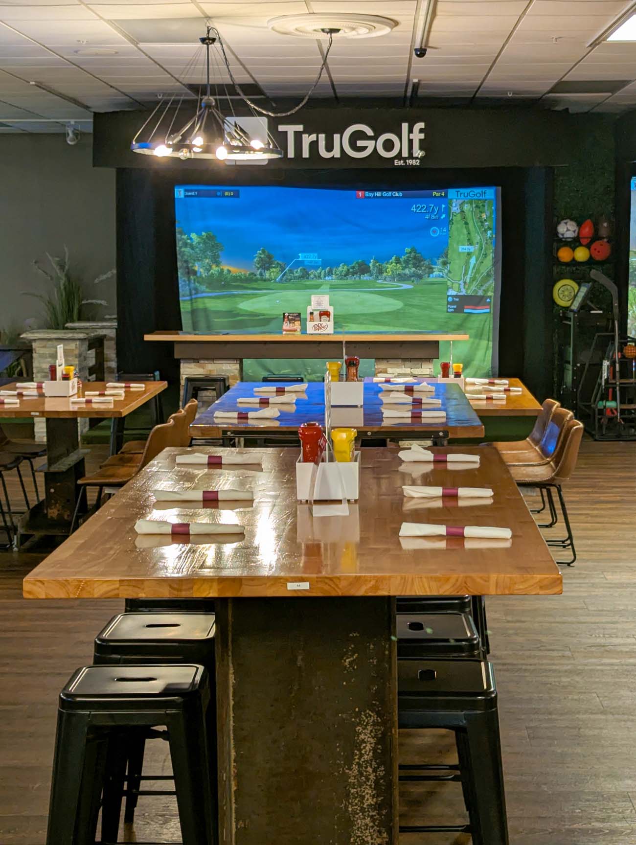 interior of Twisted Tees showing TruGolf simulator setup with large screen displaying golf course, rustic wooden tables, and modern industrial lighting