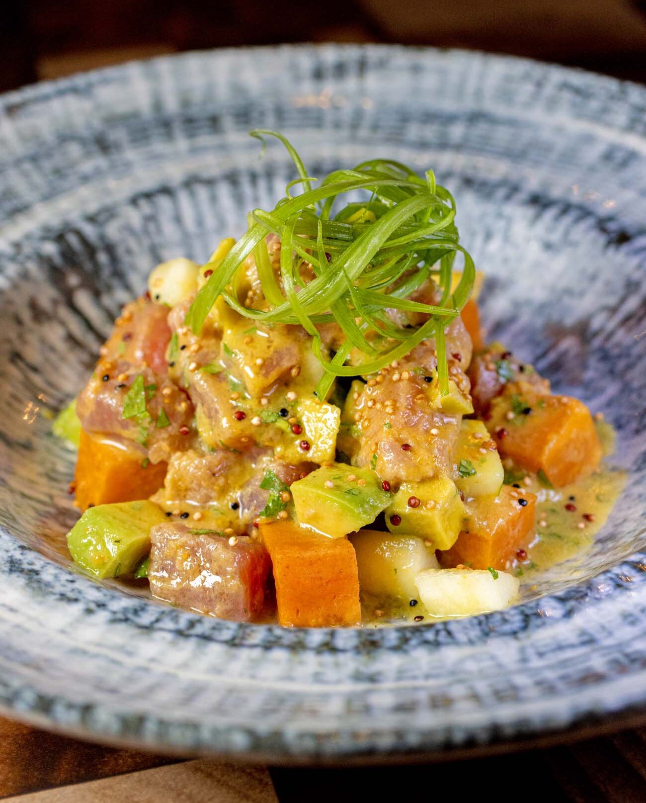 Vibrant ceviche with cubed ahi tuna, avocado, sweet potato, and microgreens garnished with sesame seeds and pink peppercorns in decorative blue and white ceramic bowl