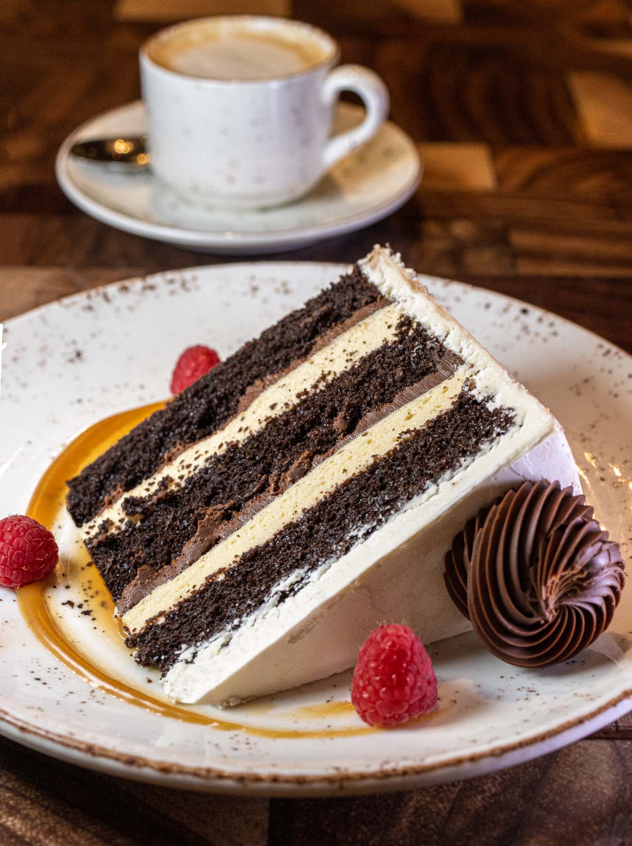 Multi-layer chocolate stout cake slice with white cream filling, garnished with fresh raspberries and chocolate decoration on speckled ceramic plate with coffee cup