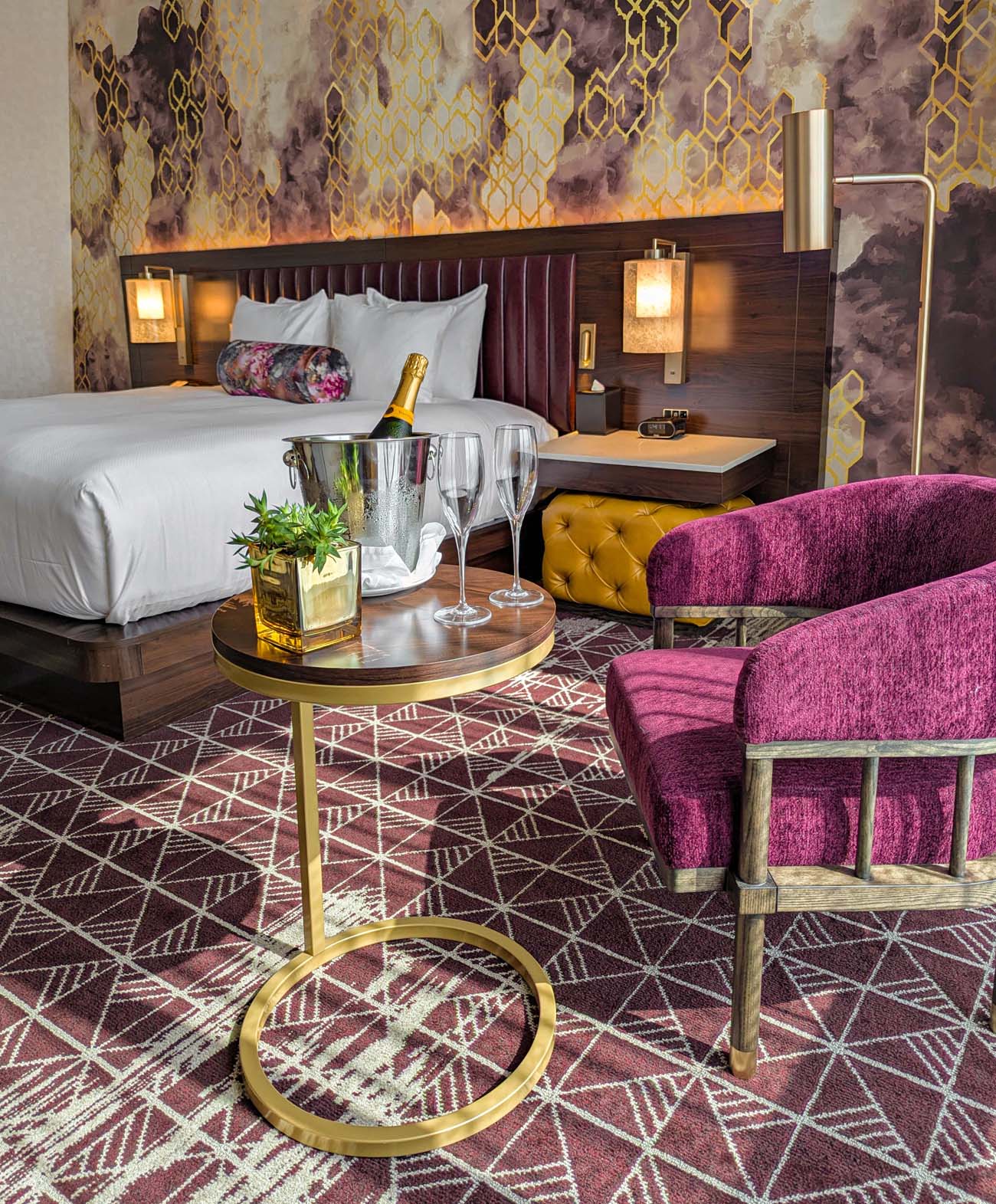 Stylish hotel room featuring purple velvet chairs, gold accent table with champagne service, rich patterned wallpaper, modern bed with burgundy headboard, and geometric carpet