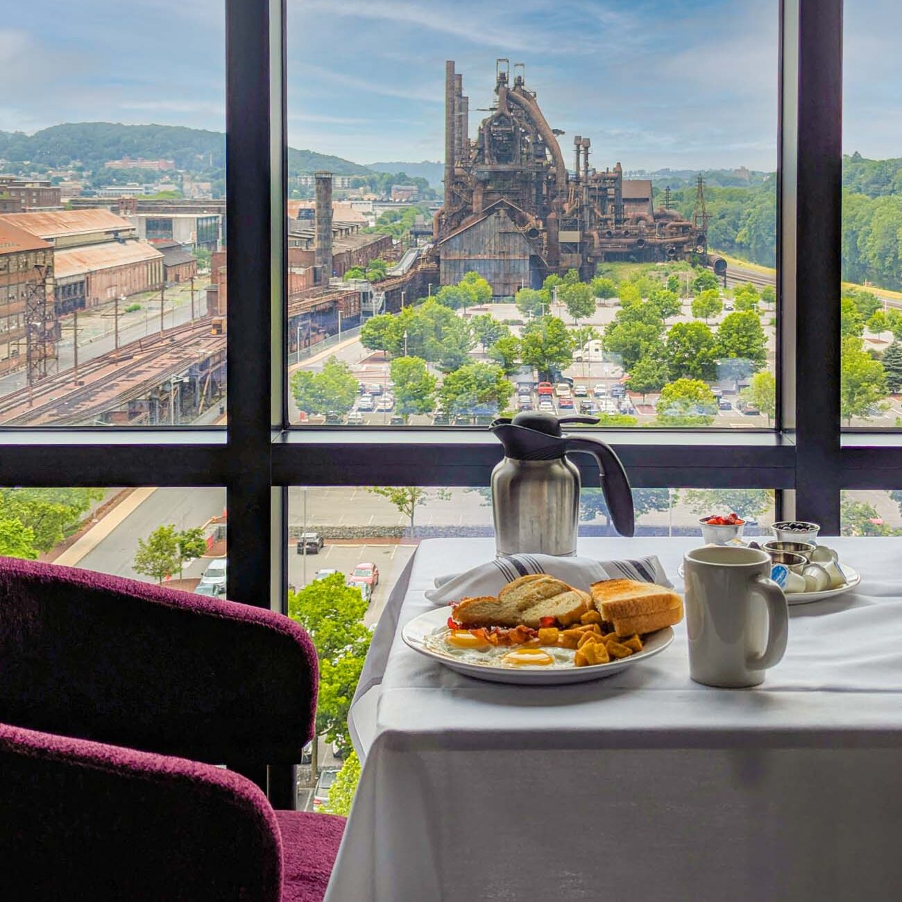 Hotel room breakfast setup by floor-to-ceiling windows overlooking historic Bethlehem Steel blast furnaces, with eggs, bacon, and coffee on white linens and purple velvet chair