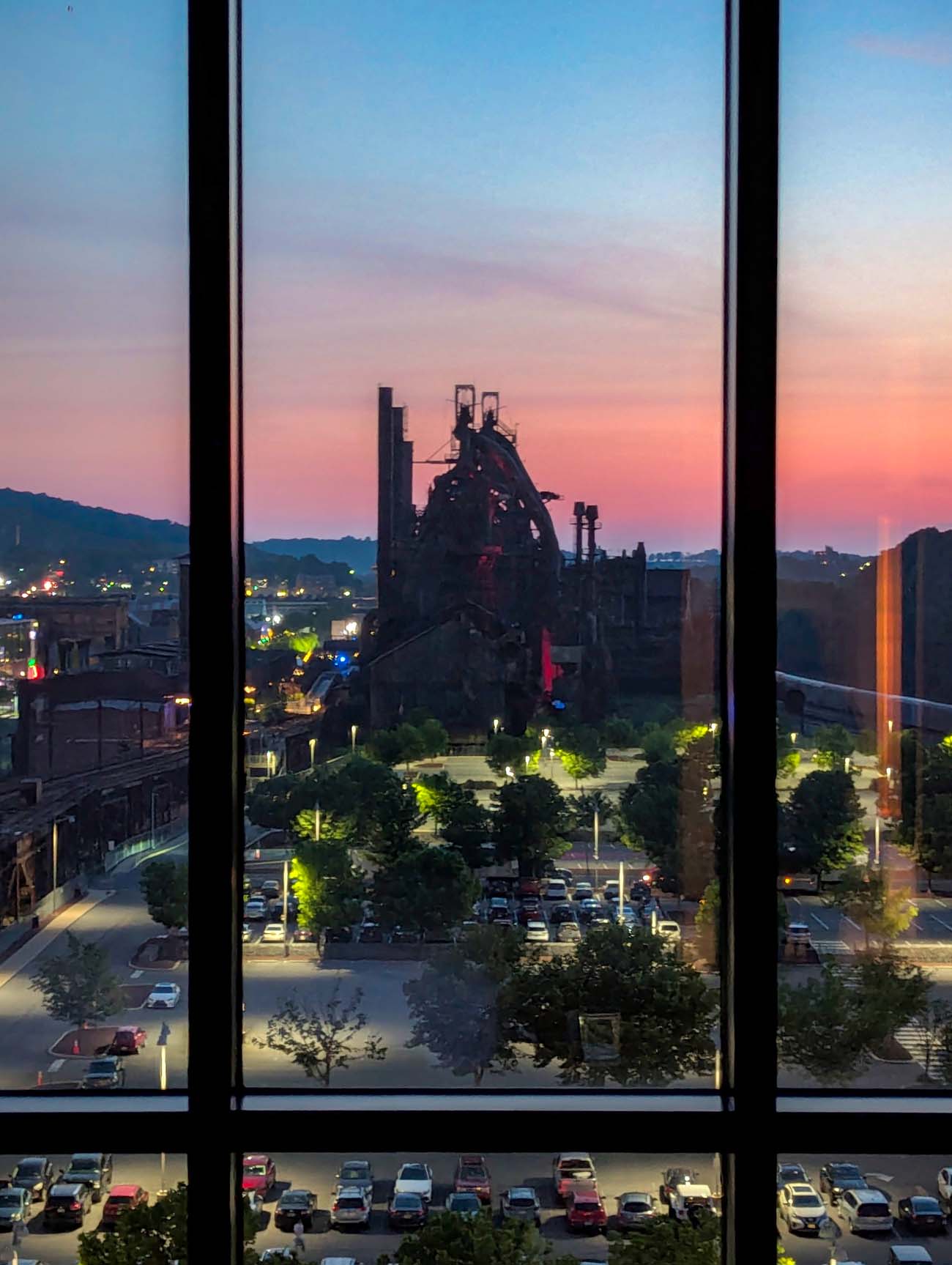 Evening view through hotel room windows showing historic steel mill structures silhouetted against pink and purple sunset sky with parking lot and green landscape below