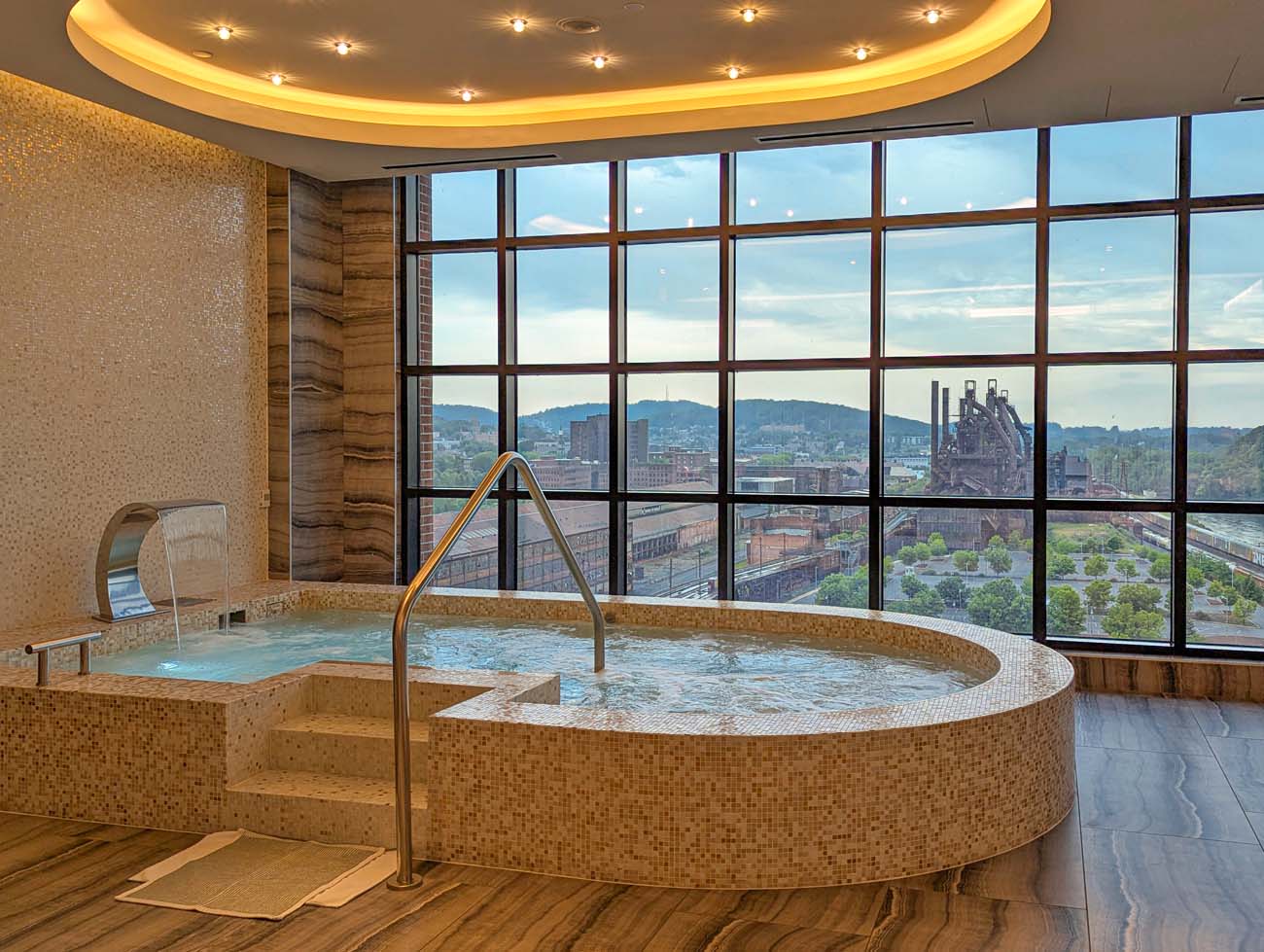 Circular mosaic-tiled vitality pool with handrails in bright spa room featuring floor-to-ceiling windows overlooking Lehigh Valley and historic SteelStacks industrial complex