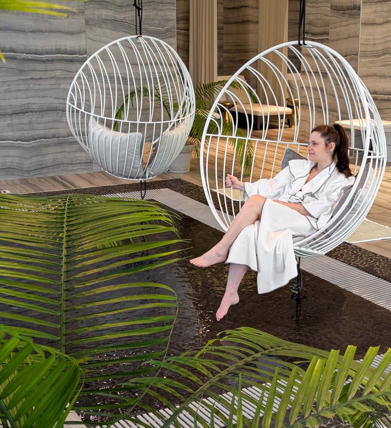 Woman in white spa robe relaxing in white wire-frame suspended swing chair over shallow wading pool, surrounded by tropical plants and natural stone spa décor