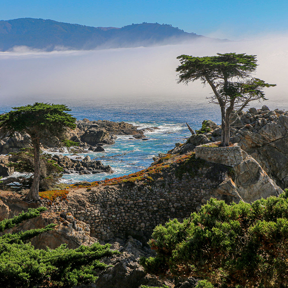 Dramatic coastal landscape of the lone cypress in pebble beach showing rocky cliffs, iconic Monterey 17 Mile drive, turquoise ocean waters, and misty mountains in the background under a partly cloudy sky