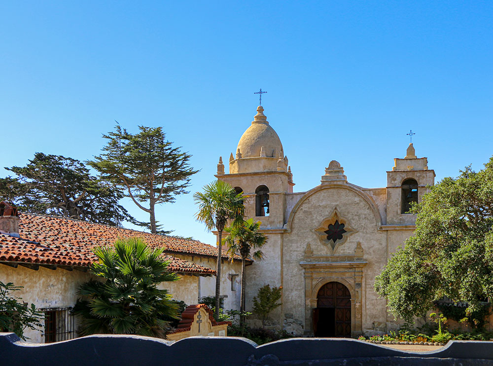 Historic Carmel Mission Basilica with distinctive Spanish colonial architecture, bell towers, palm trees, and red tile roofs against a clear blue sky