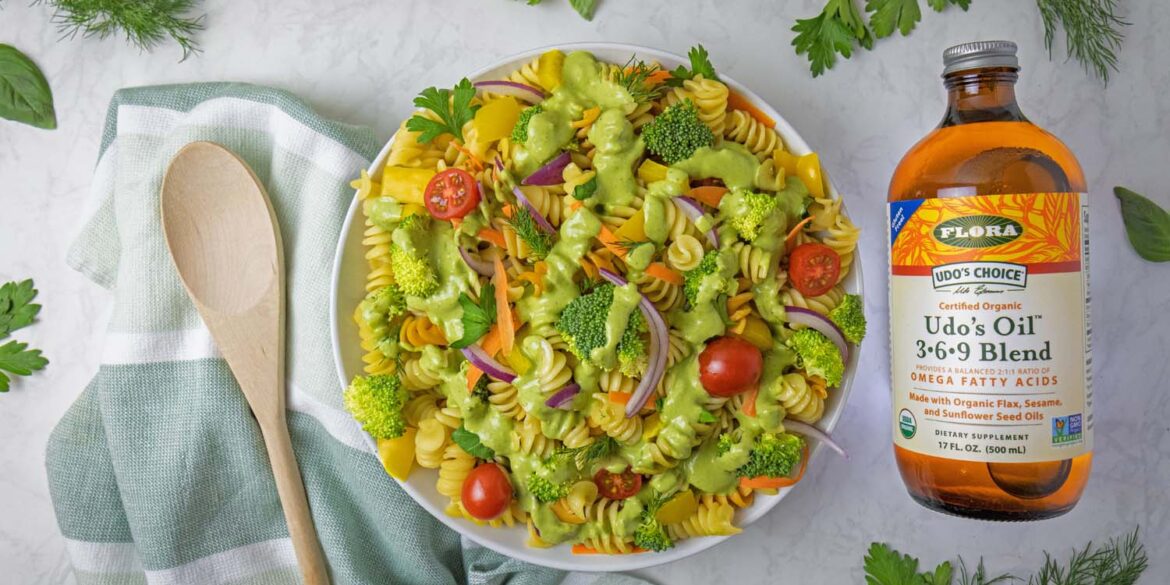 A beautifully styled overhead shot of completed Green Goddess pasta salad in a white bowl, surrounded by fresh herb sprigs including parsley, dill, and basil scattered around a white marble surface. A bottle of Udo's Oil 3-6-9 Blend sits to the right, with a wooden spoon resting on a sage green striped kitchen towel to the left. The pasta salad shows vibrant colors from the tri-color rotini, green broccoli, red cherry tomatoes, yellow bell peppers, and purple onions.