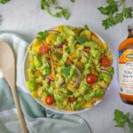 A beautifully styled overhead shot of completed Green Goddess pasta salad in a white bowl, surrounded by fresh herb sprigs including parsley, dill, and basil scattered around a white marble surface. A bottle of Udo's Oil 3-6-9 Blend sits to the right, with a wooden spoon resting on a sage green striped kitchen towel to the left. The pasta salad shows vibrant colors from the tri-color rotini, green broccoli, red cherry tomatoes, yellow bell peppers, and purple onions.