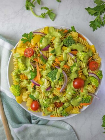 A beautifully styled overhead shot of completed Green Goddess pasta salad in a white bowl, surrounded by fresh herb sprigs including parsley, dill, and basil scattered around a white marble surface. A bottle of Udo's Oil 3-6-9 Blend sits to the right, with a wooden spoon resting on a sage green striped kitchen towel to the left. The pasta salad shows vibrant colors from the tri-color rotini, green broccoli, red cherry tomatoes, yellow bell peppers, and purple onions.