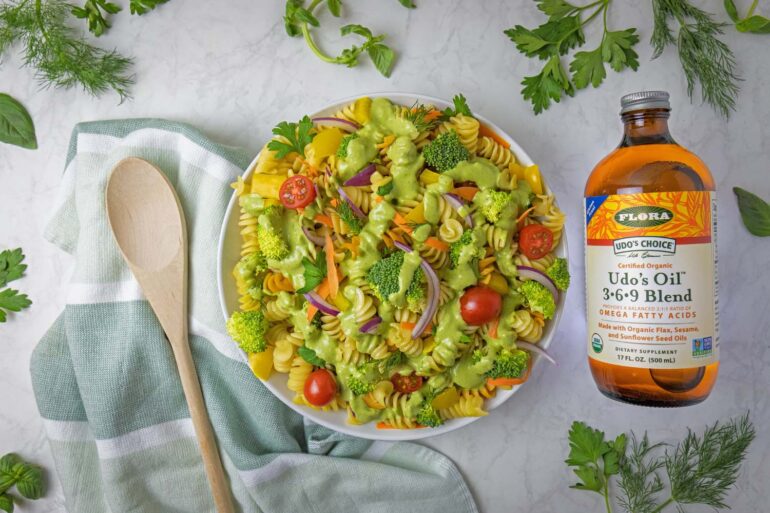 A beautifully styled overhead shot of completed Green Goddess pasta salad in a white bowl, surrounded by fresh herb sprigs including parsley, dill, and basil scattered around a white marble surface. A bottle of Udo's Oil 3-6-9 Blend sits to the right, with a wooden spoon resting on a sage green striped kitchen towel to the left. The pasta salad shows vibrant colors from the tri-color rotini, green broccoli, red cherry tomatoes, yellow bell peppers, and purple onions.