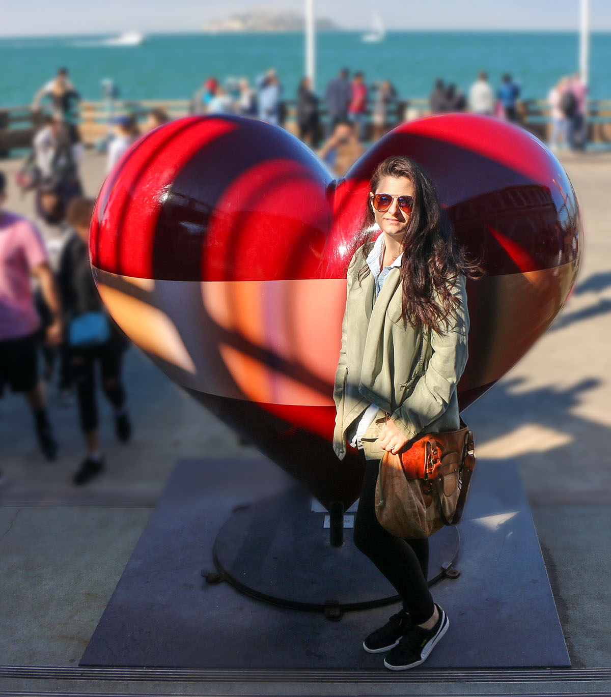 Heather from Better Living in olive green jacket and sunglasses posing next to large reflective red heart sculpture on San Francisco waterfront with people and pier visible in background