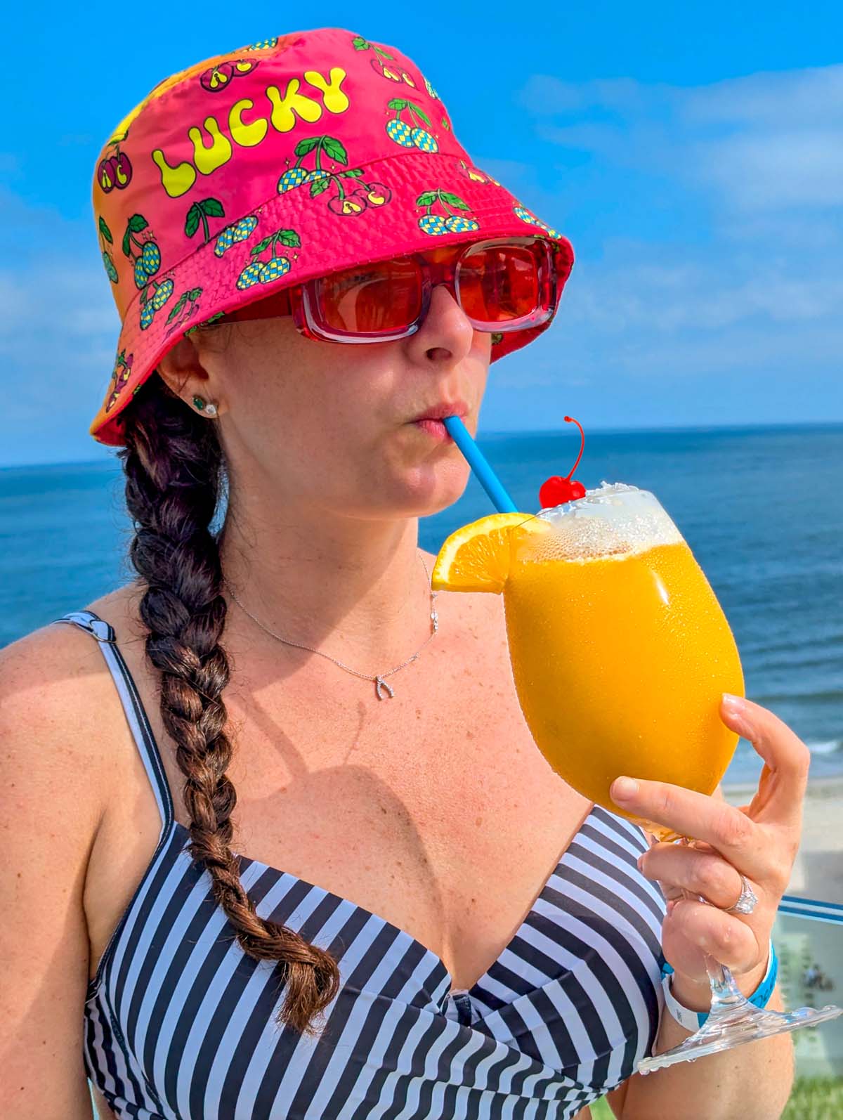 Woman in a pink "Lucky" bucket hat and striped swimsuit sipping from a yellow frozen cocktail, with ocean and blue sky in the background