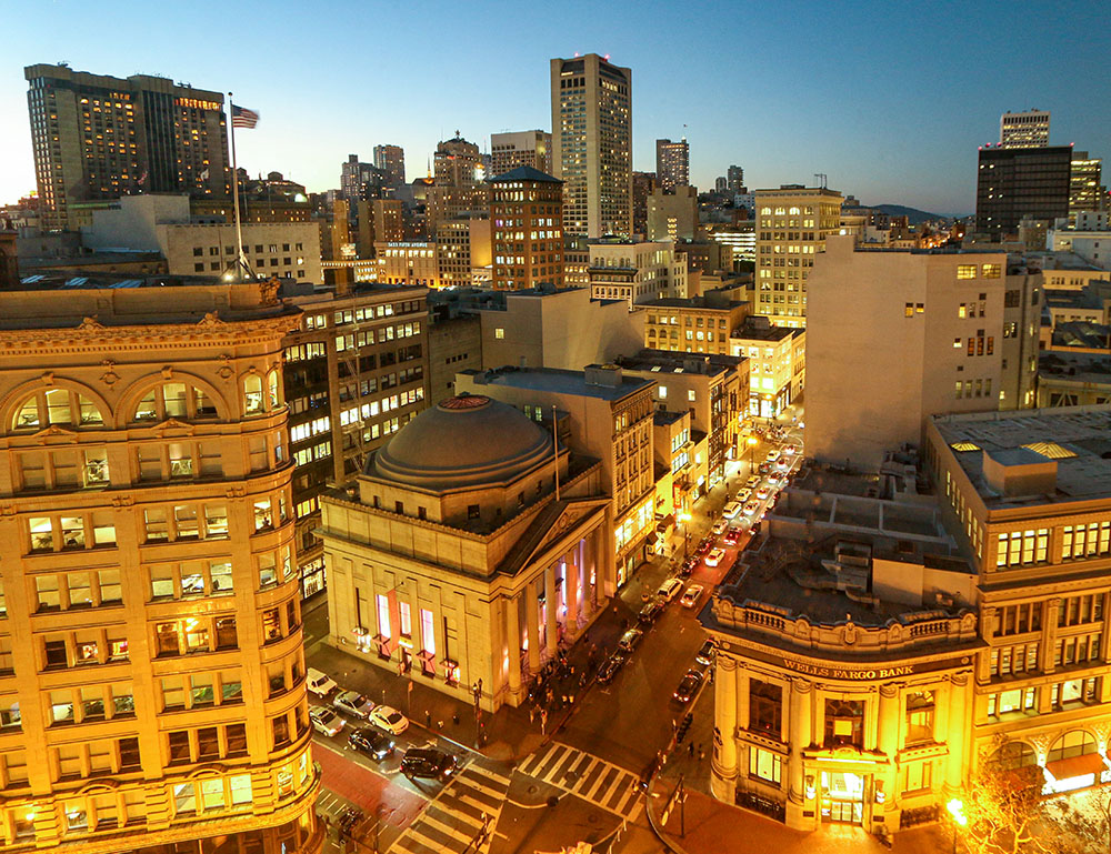 San Francisco downtown skyline at dusk showing illuminated high-rise buildings, historic architecture, busy streets with traffic, and the distinctive urban landscape from an elevated perspective