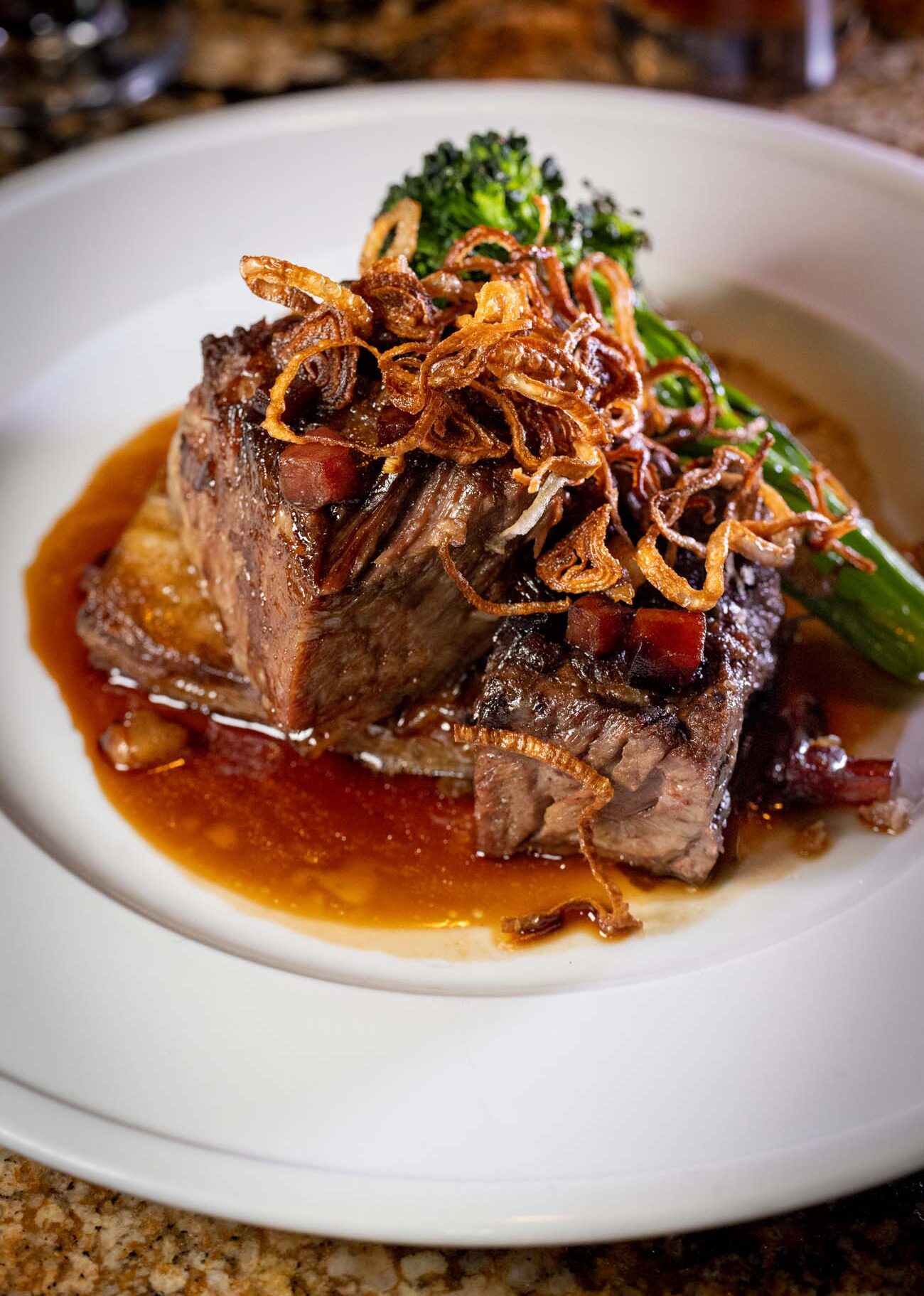 A white ceramic plate showcasing expertly plated braised beef short rib sliced to reveal tender, pink interior meat. The dish is topped with golden-brown crispy fried onions and accompanied by vibrant green vegetables including broccolini. Rich, glossy brown sauce pools around the meat, and the presentation demonstrates fine dining plating techniques on a granite countertop.