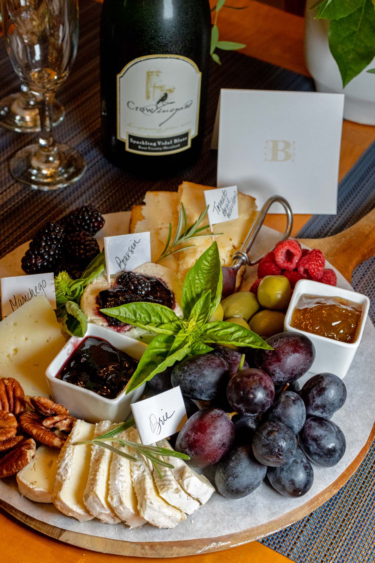 Brampton 1860 Review: A Timeless Retreat on the Eastern Shore 20 An artisan cheese and charcuterie board with Crow Sparkling Vidal Blanc wine, featuring labeled cheeses, fresh grapes, berries, and spreads arranged on a wooden platter as Brampton 1860's welcome amenity.