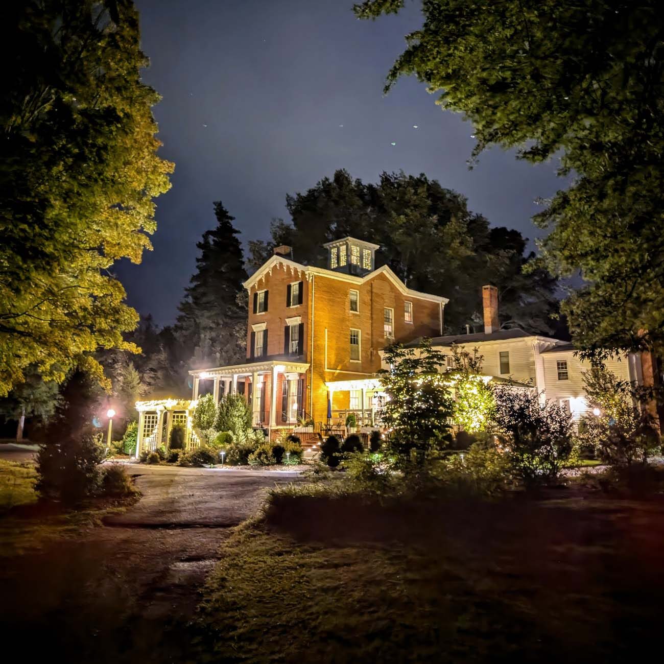Brampton 1860 Review: A Timeless Retreat on the Eastern Shore 12 Brampton 1860's Manor House illuminated at twilight with warm golden lighting highlighting the brick facade and wraparound porch, framed by mature trees under a starlit sky.
