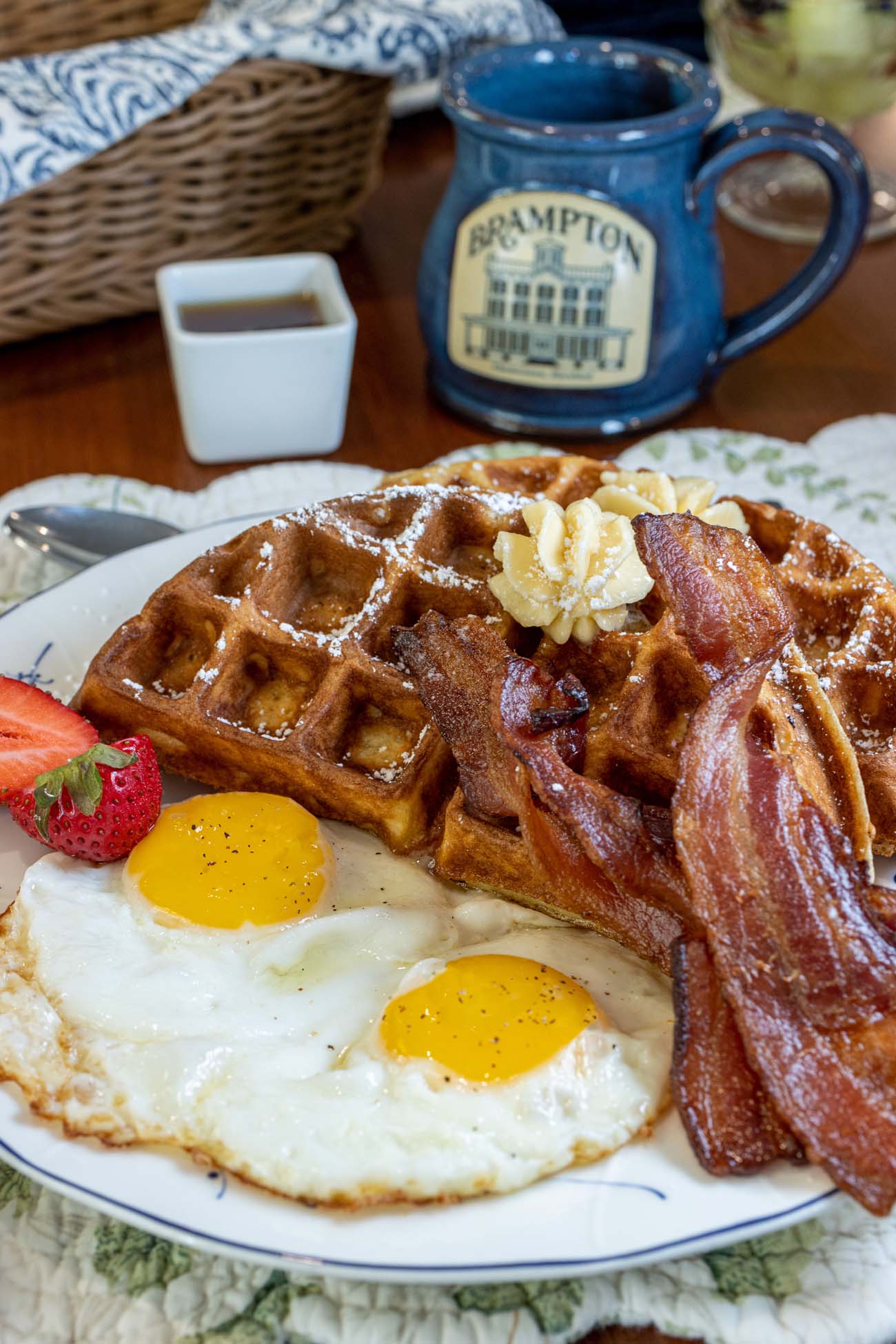 Brampton 1860 Review: A Timeless Retreat on the Eastern Shore 16 A hearty breakfast plate featuring golden waffles with butter, crispy bacon, and two sunny-side-up eggs alongside a blue ceramic Brampton 1860 commemorative mug.