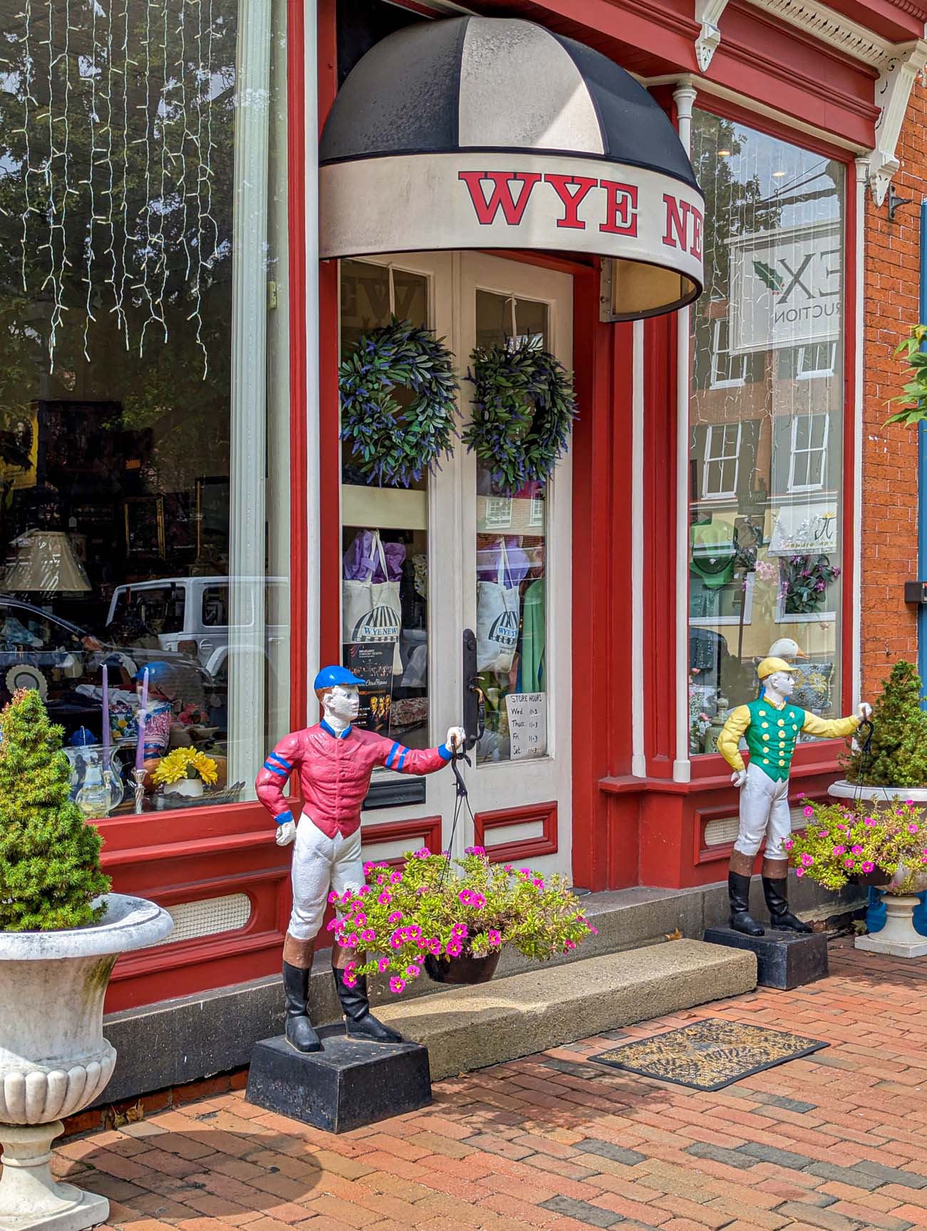 Colorful Victorian-era storefront with red and white striped trim and curved corner entrance displaying "WYE" signage. Two life-sized painted jockey statues in racing silks (one in red and blue, one in yellow and green) flank the entrance. Brick sidewalks, decorative planters with flowers, and traditional architecture create a charming small-town shopping atmosphere.