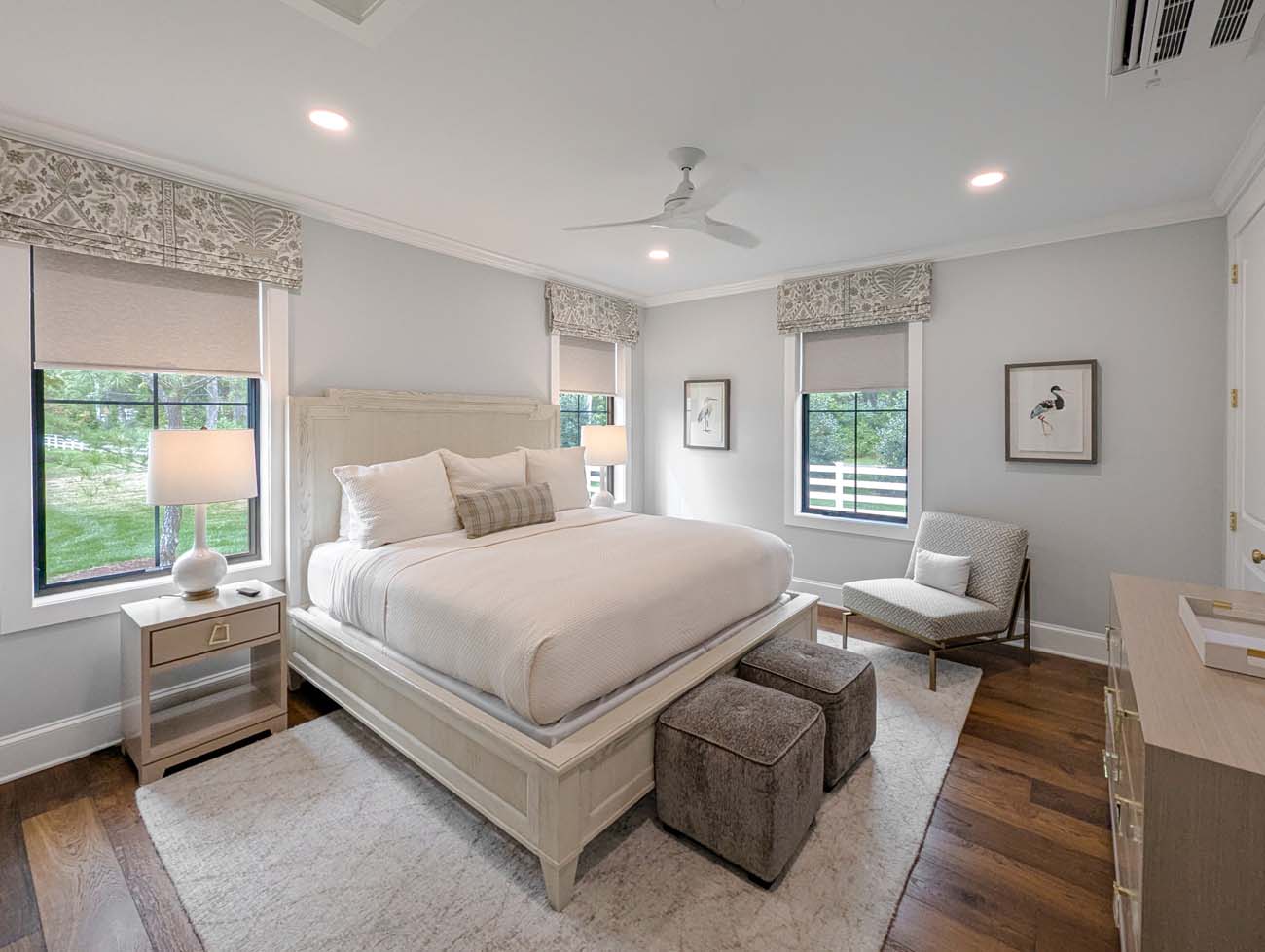 Serene master bedroom featuring a platform bed with white linens and plaid accent pillow, positioned between large windows overlooking green grounds. The room includes neutral paint, decorative Roman shades with geometric patterns, hardwood floors with area rug, modern chair, and artwork depicting waterbirds. Clean lines and natural light create a peaceful retreat atmosphere.